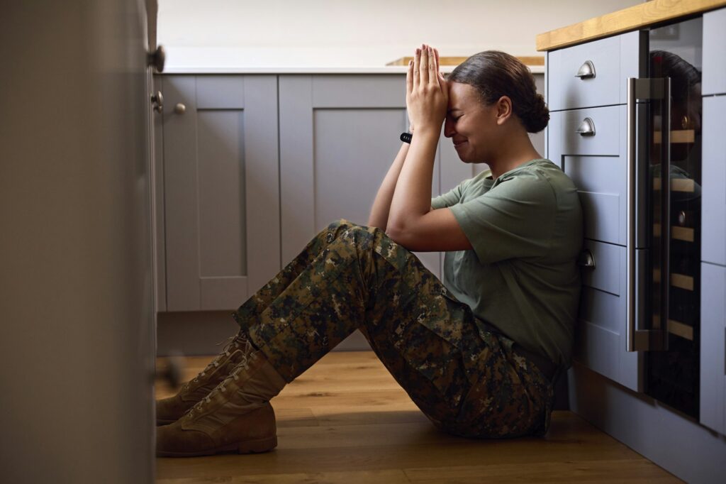 Depressed Female Soldier In Uniform Suffering With PTSD Sitting On Kitchen Floor On Home Leave